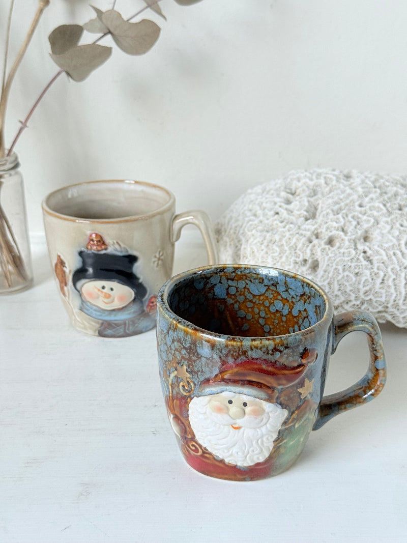 Pair of Christmas mugs with Santa and snowman designs photographed on a white surface.