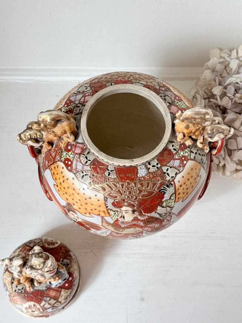 An ornate ceramic ginger jar with a matching lid, decorated with detailed hand-painted figures and animals in red, gold, and white, set against a white background.