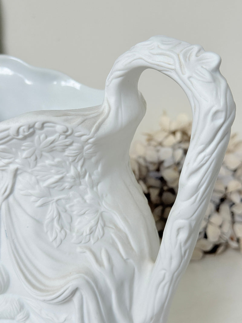 A close-up of a white ceramic pitcher with intricate embossed floral and leaf patterns, featuring a curved handle and a soft-focus decorative background.