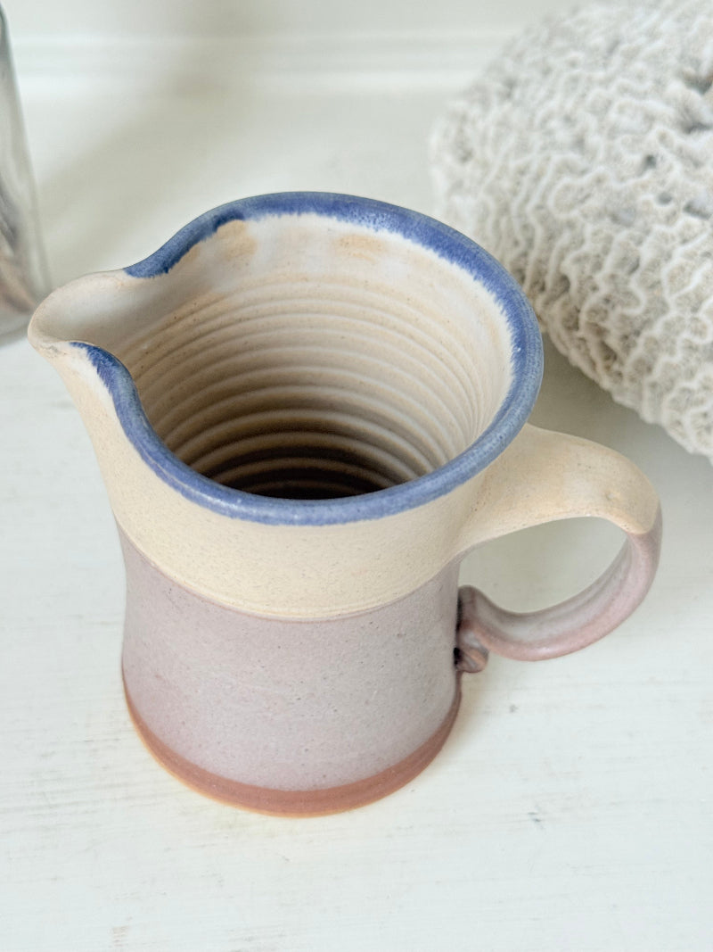 Studio pottery milk jug in cream and lavender glaze with impressed initials, photographed on a white surface.