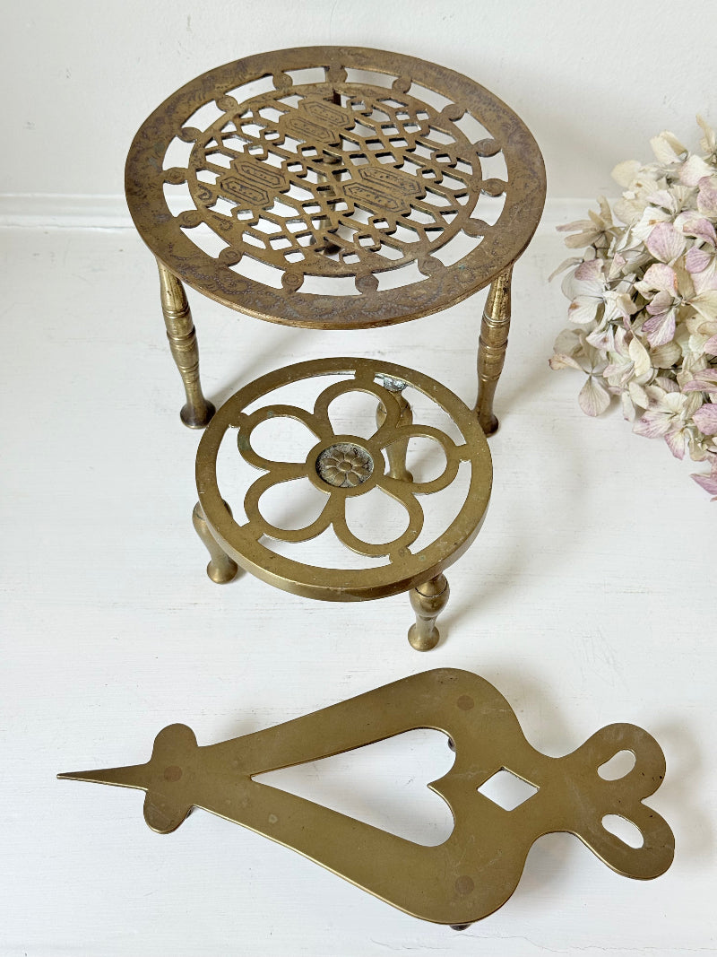 Vintage brass trivet trio with ornate cutwork designs and aged patina, photographed on a white surface