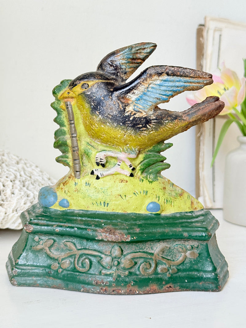 A vintage hand-painted cast iron bird feeder and money box featuring a robin holding a penny, set against a softly blurred background with a white vase and decorative items.