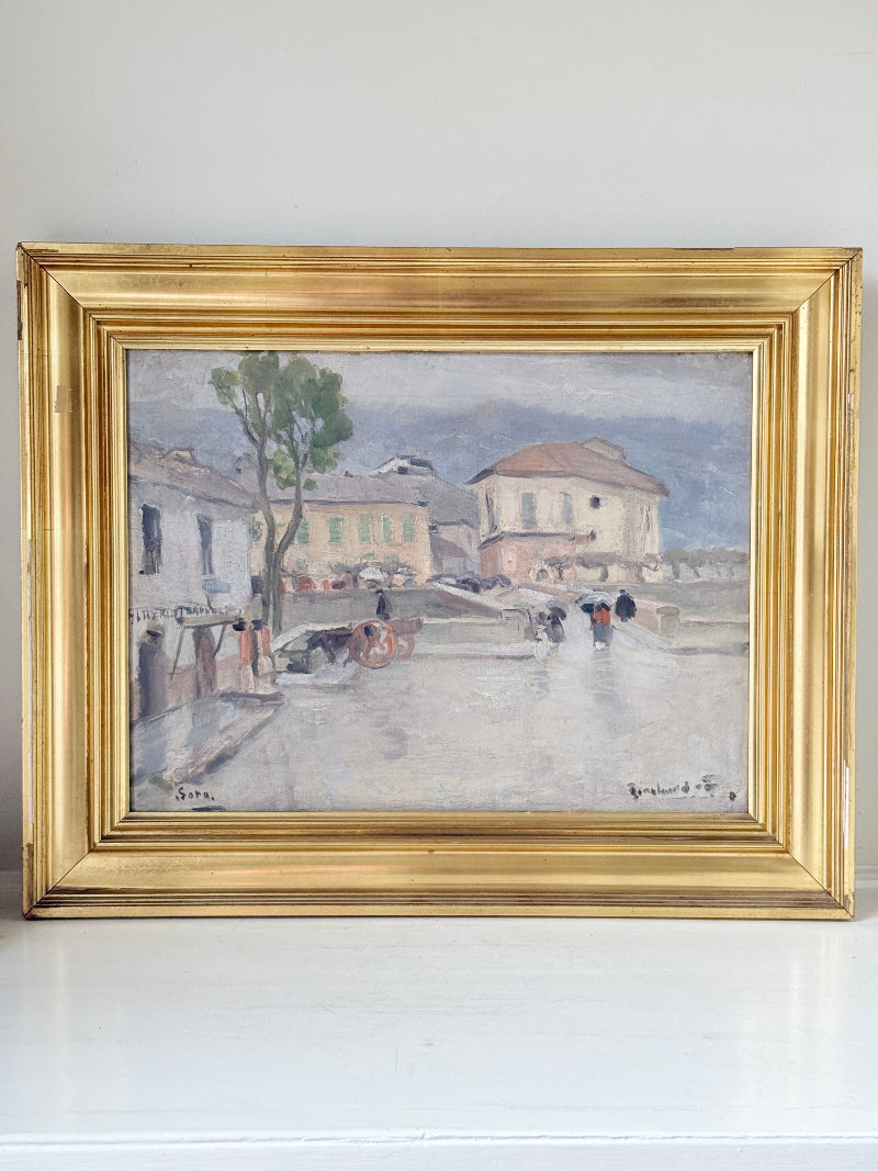 A framed impressionist landscape painting depicting a street scene with buildings, a tree, and figures, housed in a wide ornate gold frame and resting on a white surface against a white wall.