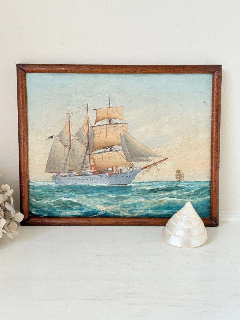 A framed vintage oil painting of a tall sailing ship on blue waves, housed in a dark wood frame and displayed on a white surface with decorative props.