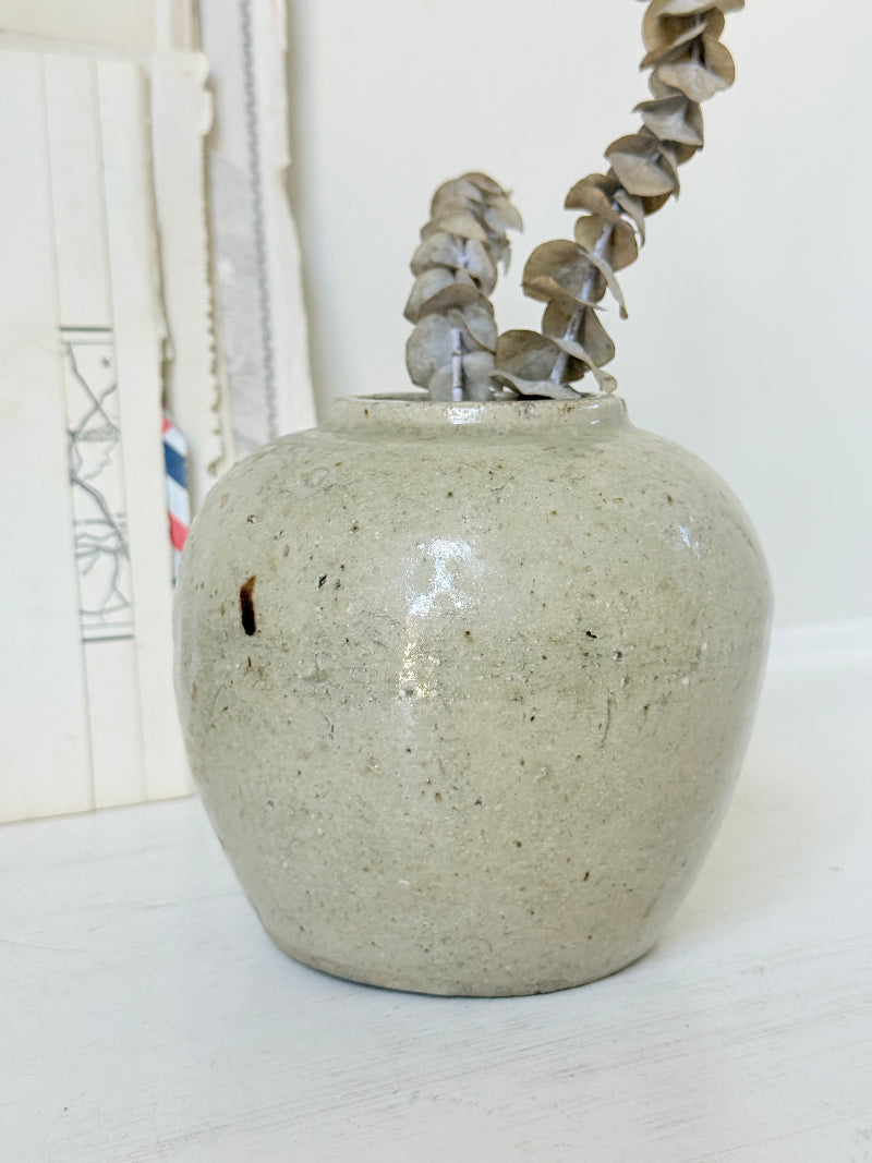 A small handmade ceramic vase with a speckled light gray-green glaze, holding a dried eucalyptus branch, set against a soft white background with blurred books on the left.