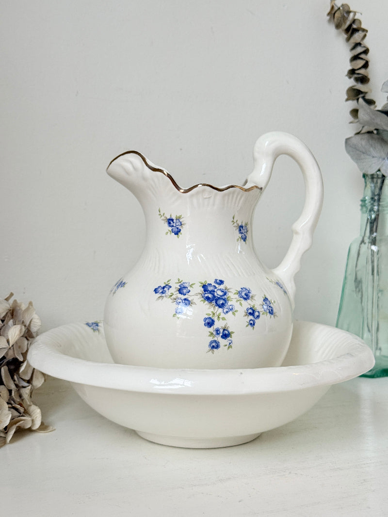A white ceramic pitcher with blue floral patterns and a gold-trimmed spout, displayed on a matching white footed bowl against a light gray background.
