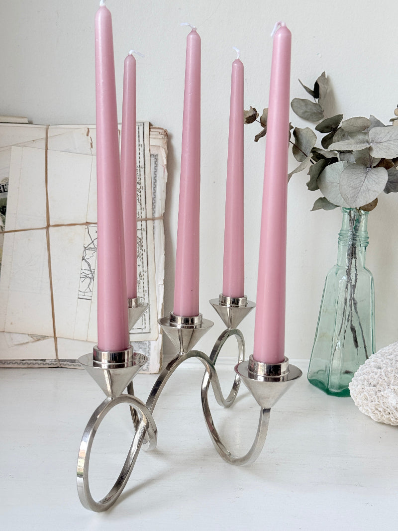 Silver tone five light candelabra with a sculptural looped base holding pink candles, photographed on a white surface