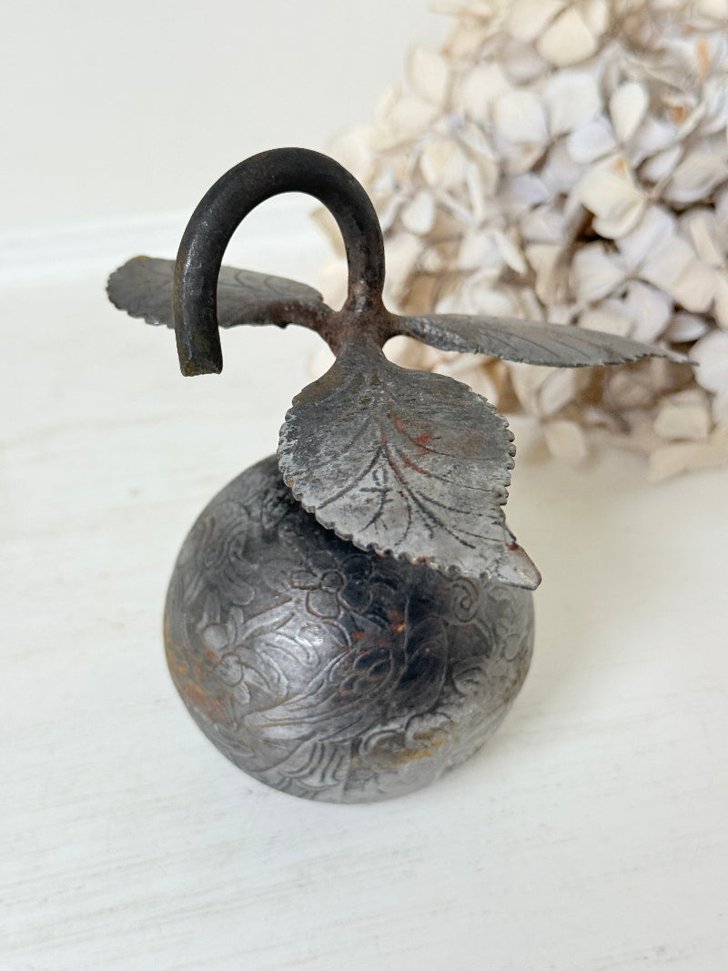 A vintage-style metal decorative bell with an engraved floral pattern, featuring a leaf-shaped handle and a curved hook at the top, shown on a light surface with a blurred dried floral background.