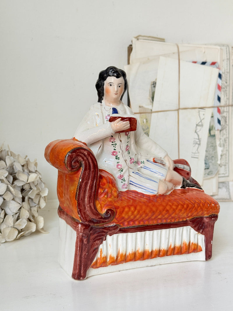 A vintage-style ceramic figurine of a man reclining on an orange chaise lounge, holding a red book. The figurine is dressed in a white floral jacket with blue striped shorts.