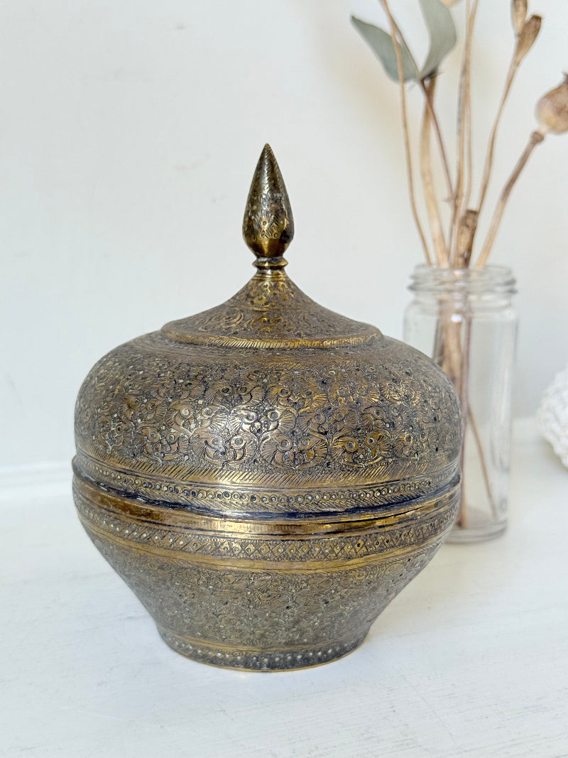 Indian Benares brass lidded jar with pointed finial and darkened interior, photographed on a white surface