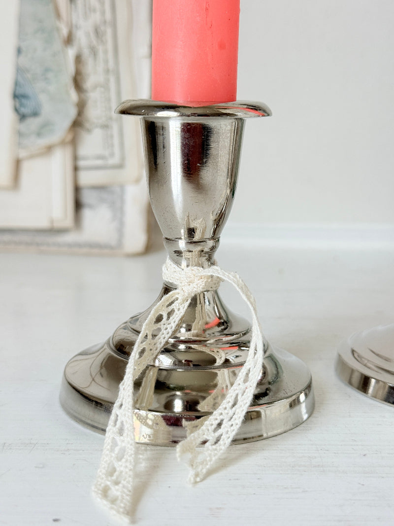 Pair of silver tone candlesticks with urn sconces and round stepped bases holding coral candles, photographed on a white surface