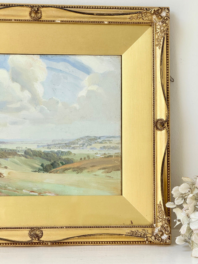 Framed oil painting of a rolling countryside landscape under a dramatic cloud-filled sky in an ornate gilt frame with wide mount.