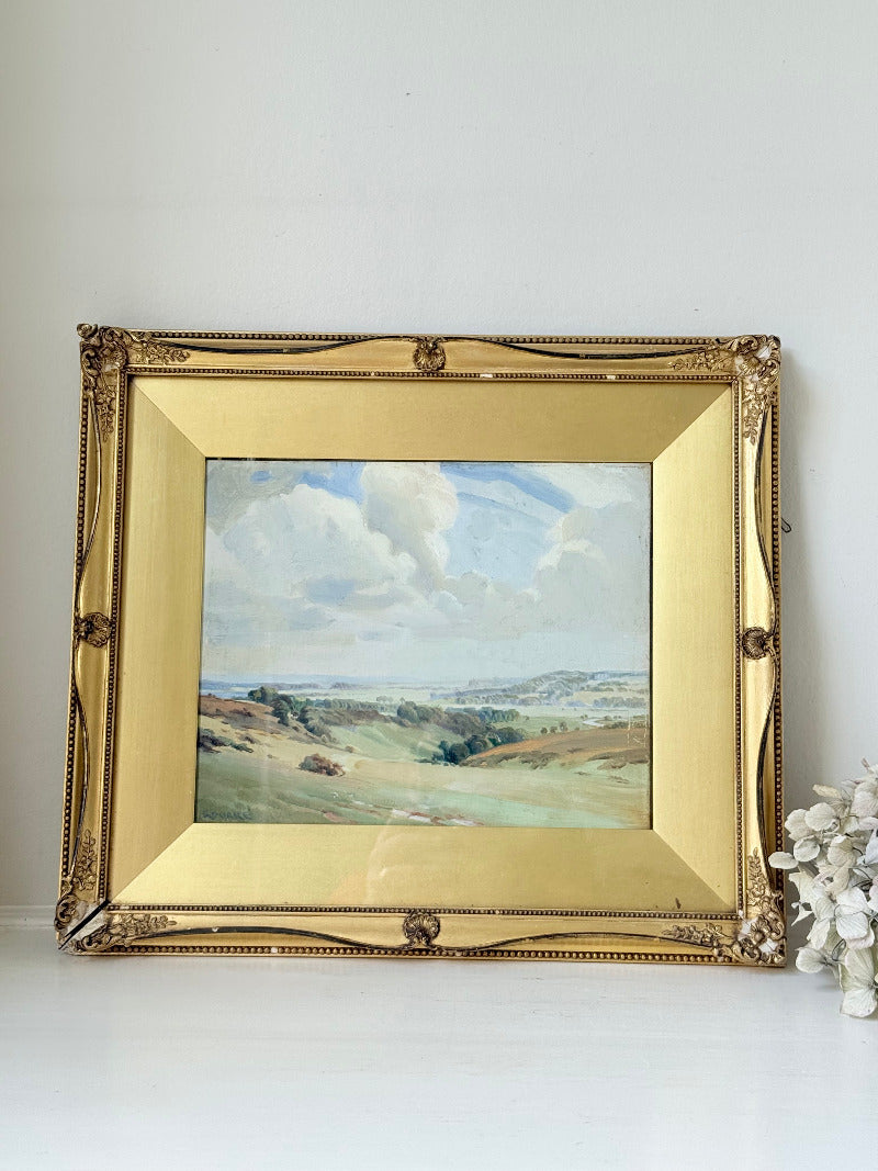 Framed oil painting of a rolling countryside landscape under a dramatic cloud-filled sky in an ornate gilt frame with wide mount.
