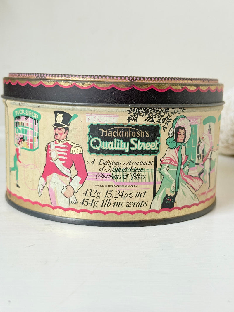 Vintage Quality Street tin from 1986 with colourful illustrated street scenes and pastel design, photographed on a white surface.