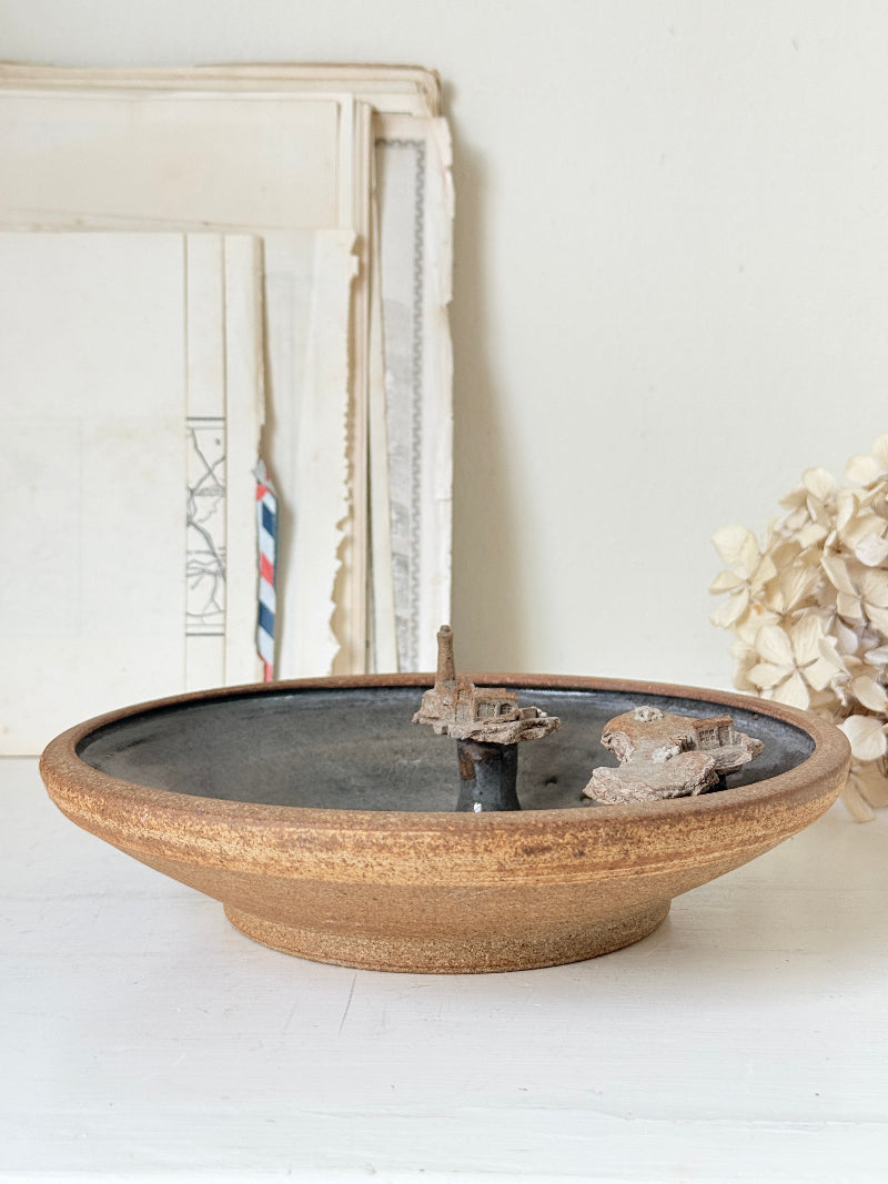 A rustic ceramic bowl with a wide, textured tan-brown exterior and a dark charcoal-gray glazed interior, styled on a white surface with vintage frames and dried flowers in the background.