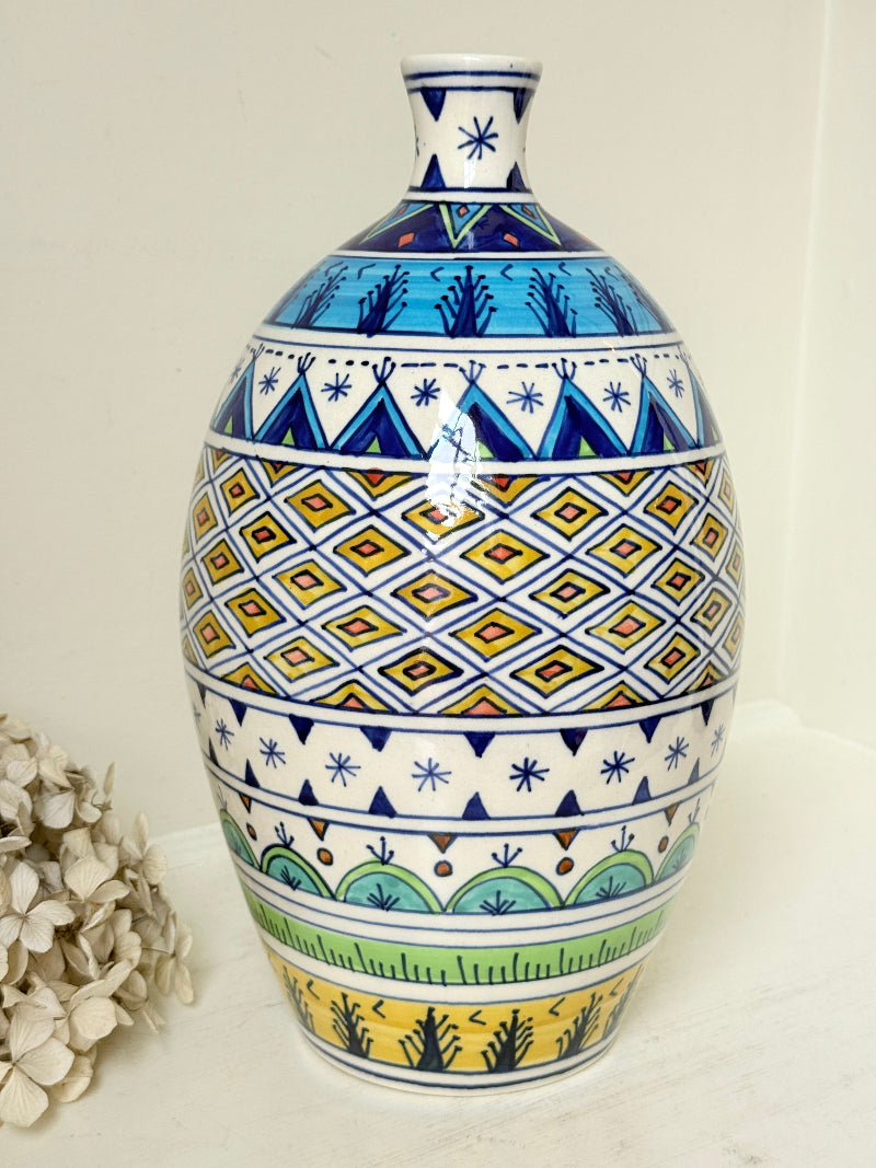 A ceramic vase with a Mediterranean or Italian Polynesian style pattern, featuring blue, yellow, green, and white geometric designs, displayed on a white surface next to dried flowers.