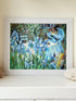 Framed impressionist-style oil painting of blue irises and garden scene, photographed on a white surface