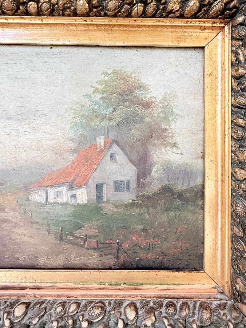 An antique oil painting of a small white cottage with a red-orange roof, set in a grassy field with a large tree behind it, housed in an ornate carved gold frame.