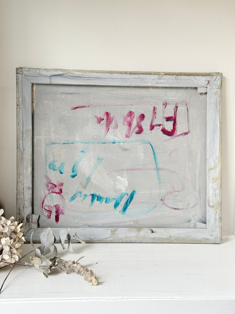 A distressed, vintage-style square frame with a weathered gray and white wood border, containing abstract scribbled artwork in pink and light blue on a translucent or off-white center. The frame is displayed on a white shelf against a beige wall, styled with dried flowers in the foreground.