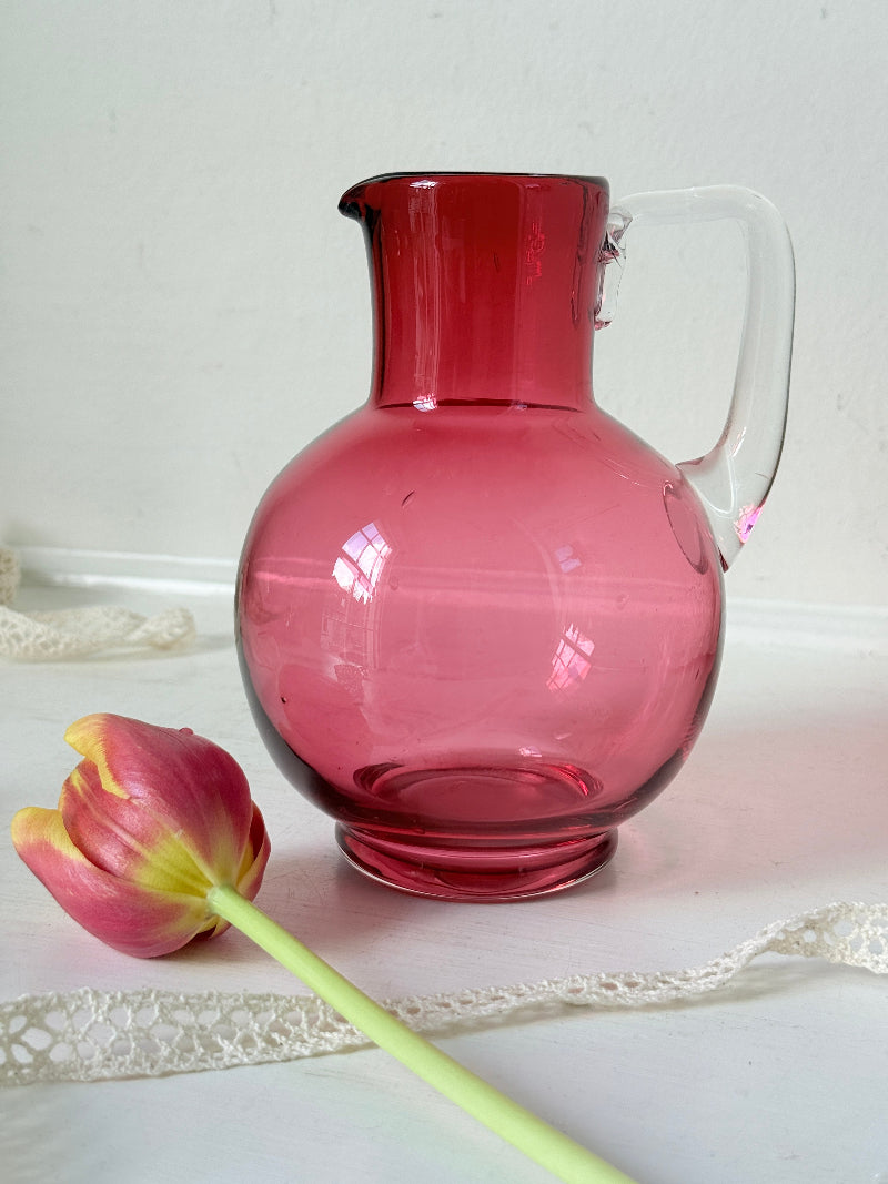 Vintage cranberry glass jug with clear handle photographed on a white surface, bulbous pink body with flared pouring rim