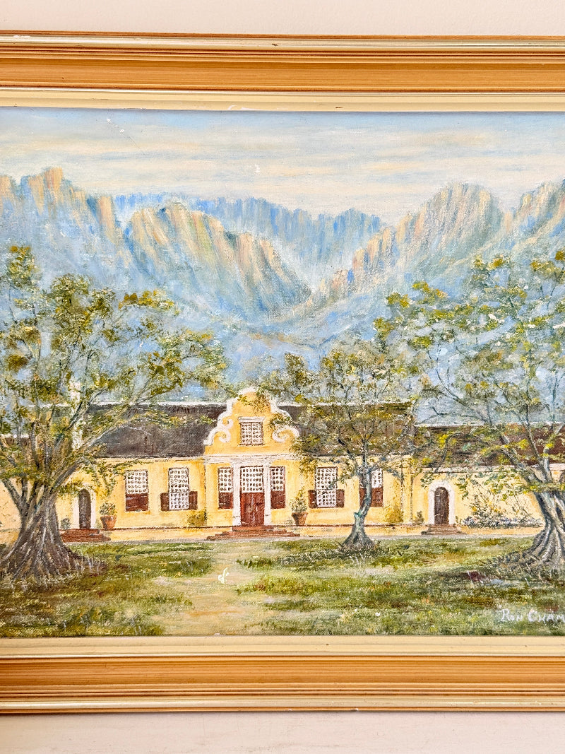 A framed oil painting depicting a yellow colonial-style building with red doors, surrounded by green trees, set against a backdrop of blue and gray mountains under a pale sky. The painting is housed in a wide, ornate gold-toned frame.