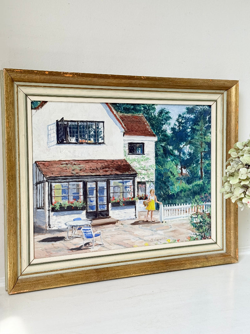 Oil on board painting of a girl in a yellow dress outside a cottage with picket fence, framed, photographed on a white surface