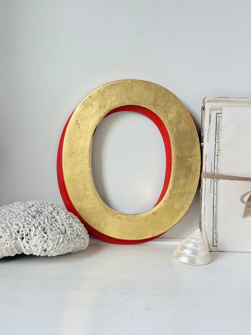 A large decorative letter O wall piece with a textured gold finish and a red painted inner border, styled on a white surface with a white crochet pillow, a small white shell, and a vintage-style book.