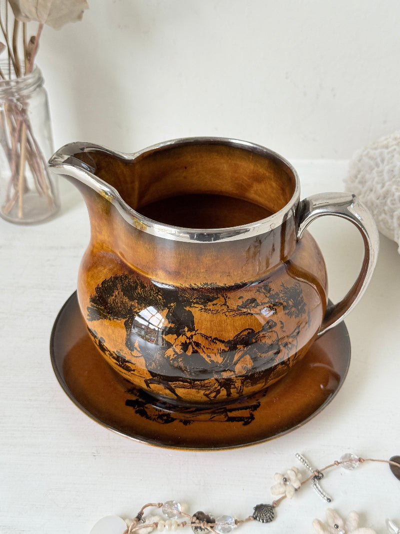 Antique Ridgways stoneware creamer jug and saucer photographed on a white surface in amber treacle glaze with black coaching scenes and silver lustre rim