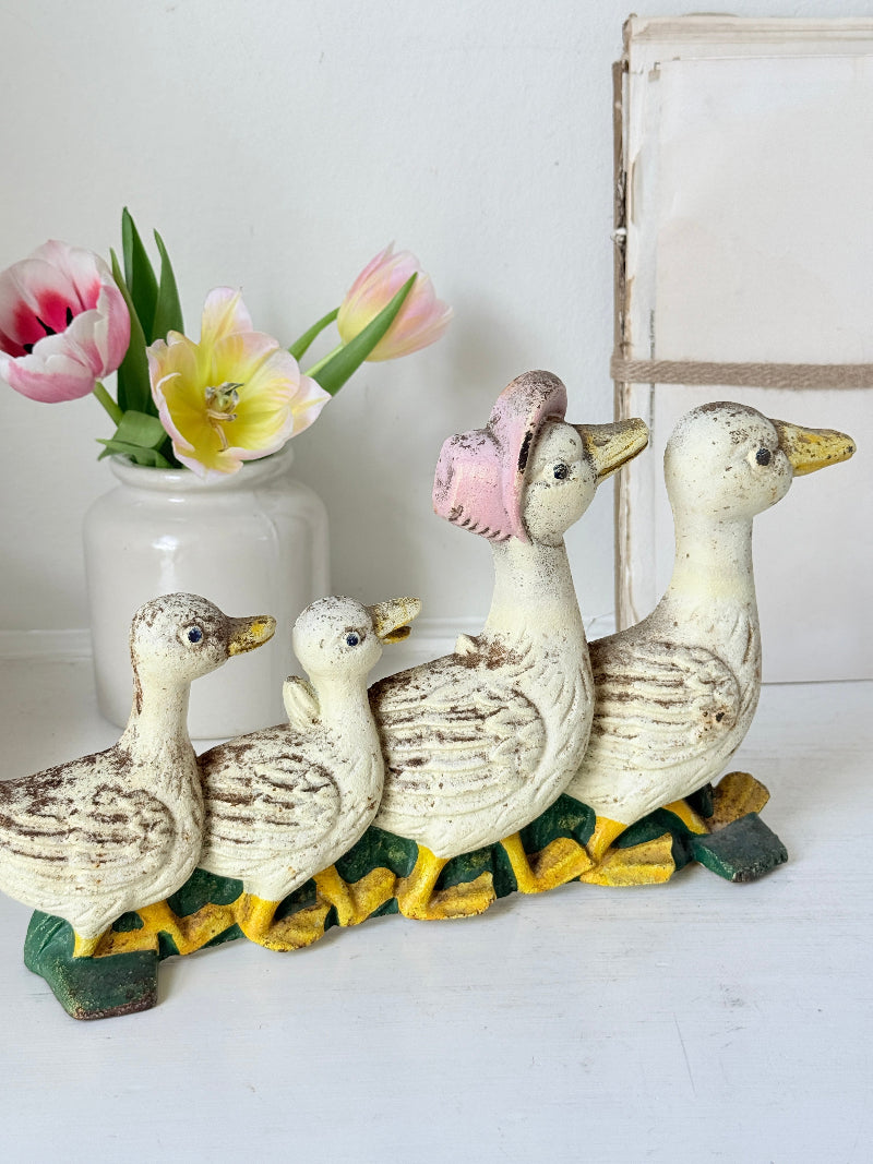 A vintage-style painted family of four ducks figurine in cream, brown, yellow, and green, displayed on a white surface next to a white ceramic vase with pink and yellow tulips.