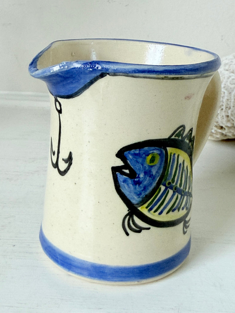 Vintage studio pottery jug with hand painted blue and green fish design on cream clay, photographed on a white surface.