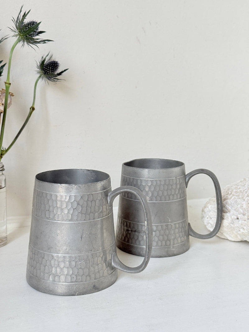 Pair of vintage pewter tankards with handles and textured detail, photographed on a white surface