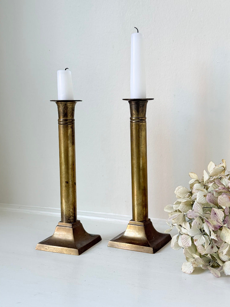 Pair of vintage brass candlesticks with square bases and white taper candles, styled on a white surface beside dried hydrangeas.