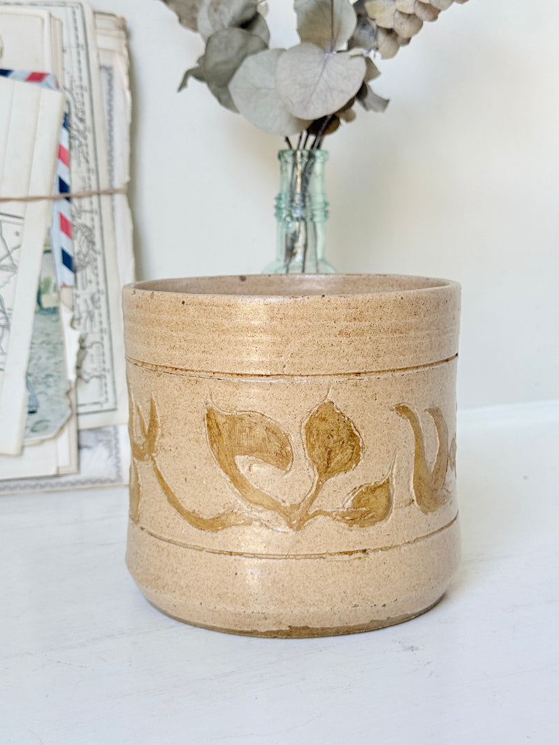 A handmade ceramic planter in a sandy beige stoneware finish with a rustic brown leaf pattern, styled on a white surface with dried flowers in a glass vase and vintage postcards in the background.