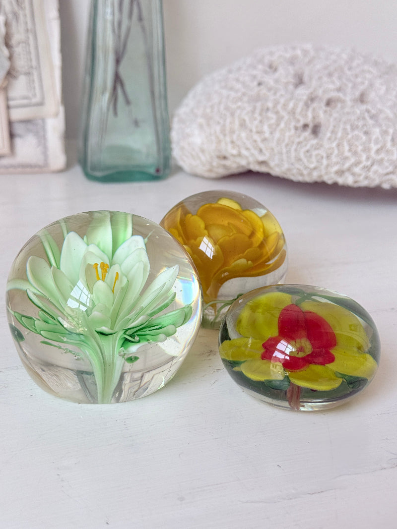 Trio of mixed flower glass paperweights in green, yellow and red, graduated in size, photographed on a white surface