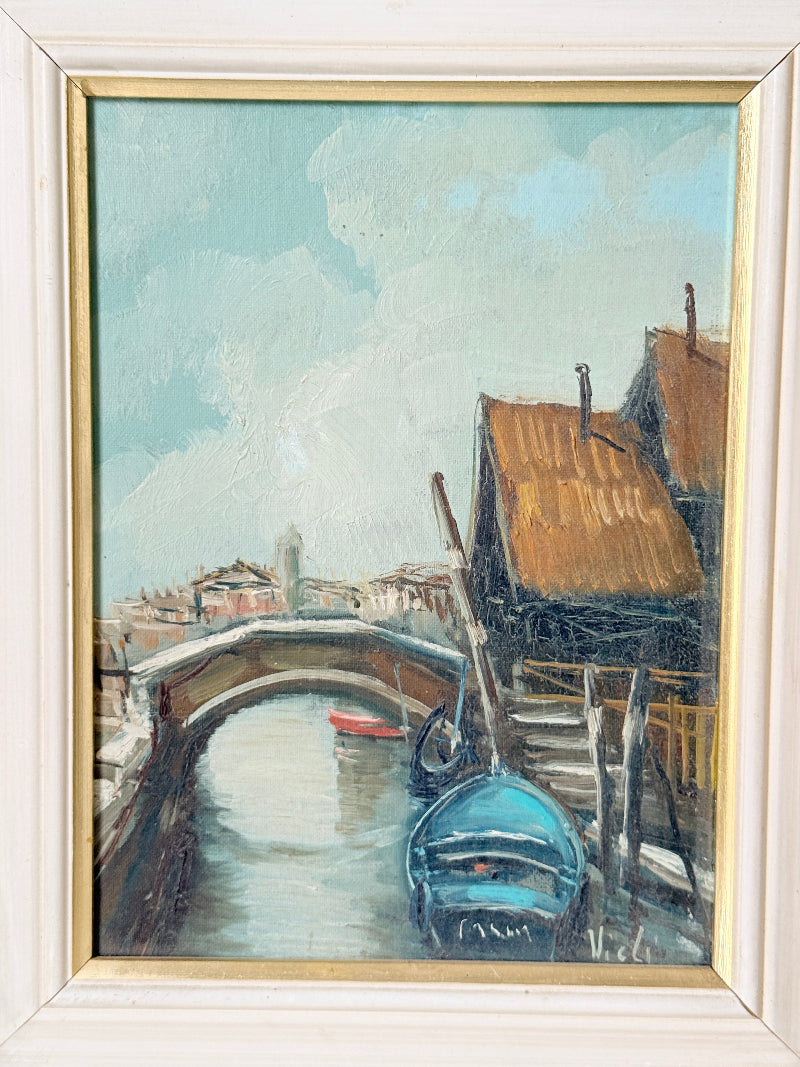 A framed oil painting depicting a Venetian canal scene with an arched bridge, rustic buildings with orange-brown roofs, and a blue gondola in the foreground, set within a light-colored wooden frame with a gold inner border.