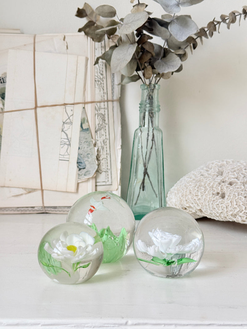 Trio of white flower glass paperweights in graduated sizes with green leaves, photographed on a white surface
