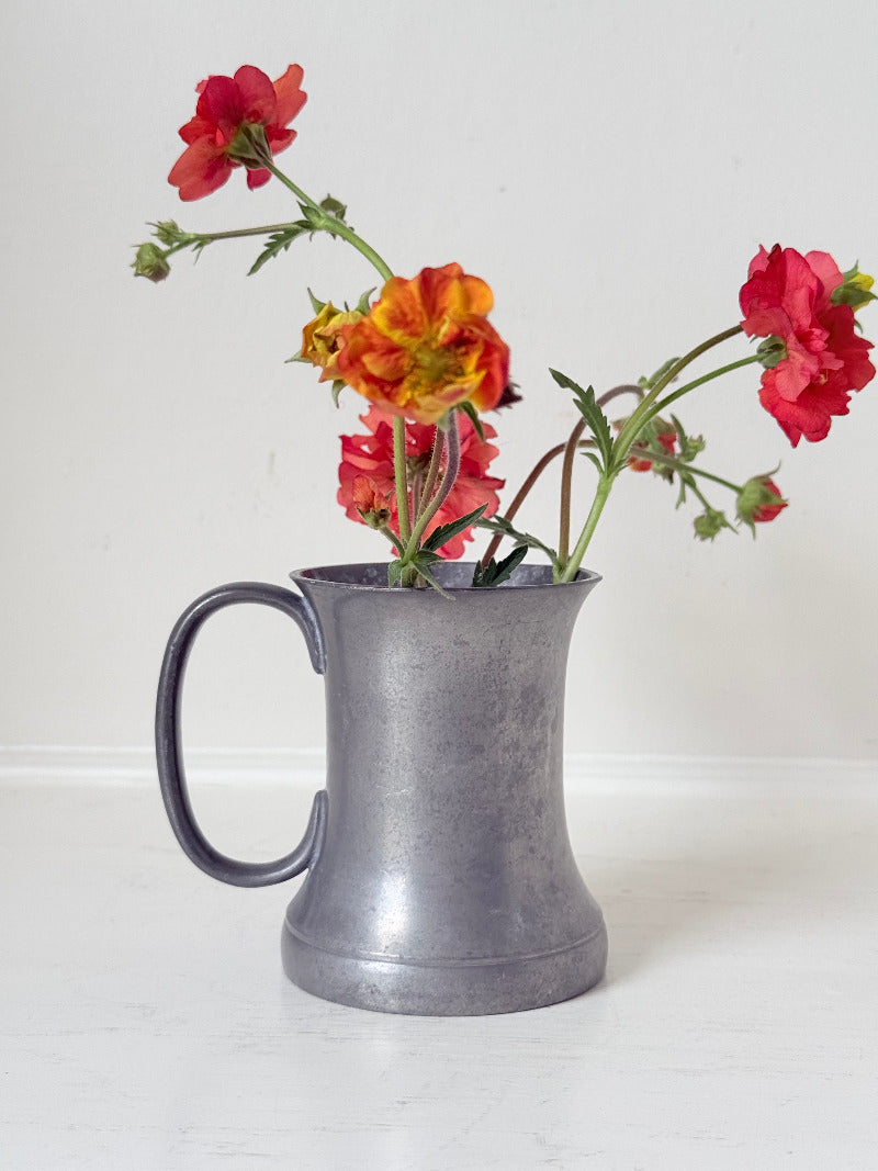 vintage pewter tankard with crown and rose mark and handle, styled with red and orange flowers on white surface