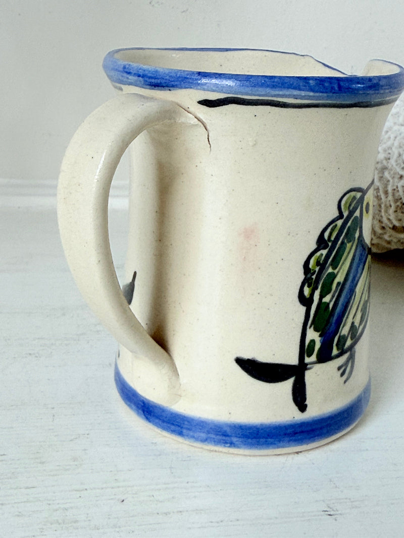 Vintage studio pottery jug with hand painted blue and green fish design on cream clay, photographed on a white surface.
