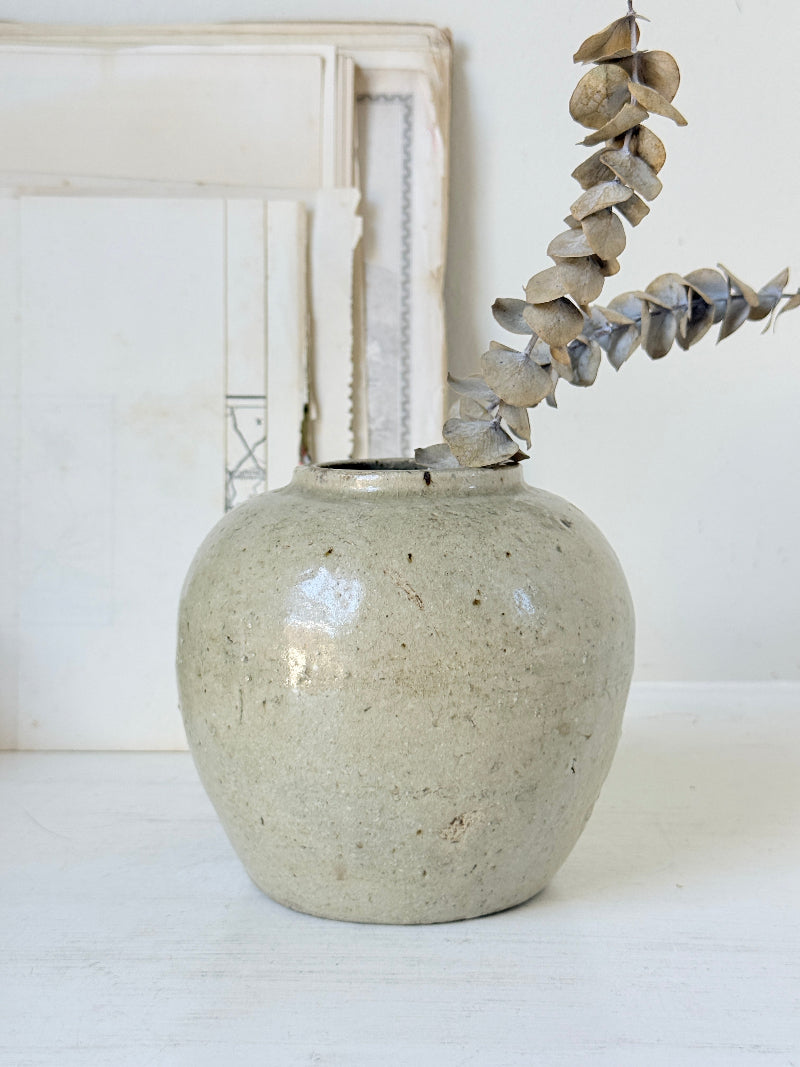 A handmade ceramic vase with a speckled, matte light gray-beige glaze, holding a dried eucalyptus branch, set against a rustic white background with a distressed wooden frame.