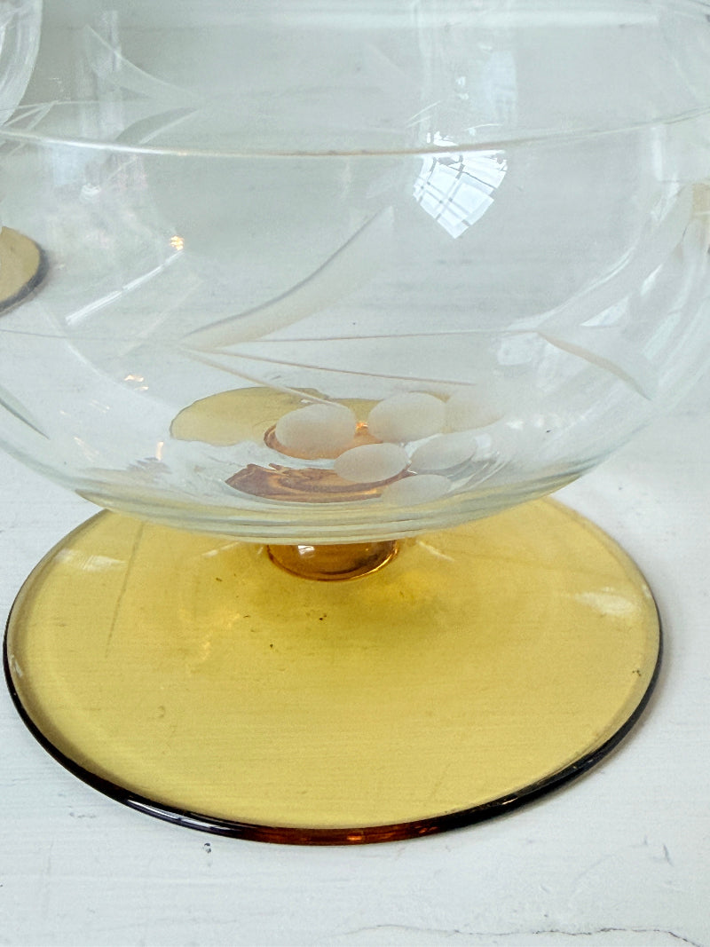 A clear glass bowl with a wide, round amber-yellow pedestal base, photographed on a white surface.