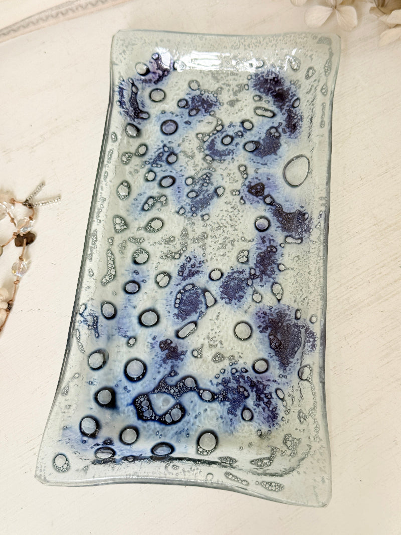 A rectangular glass tray with a wavy edge, decorated with blue and purple bubble patterns on a clear glass base, displayed on a light-colored wooden surface.