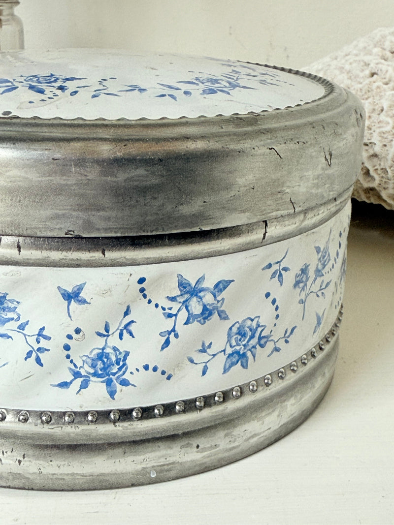 Vintage round shortbread tin with blue floral pattern on silver background, photographed on a white surface.