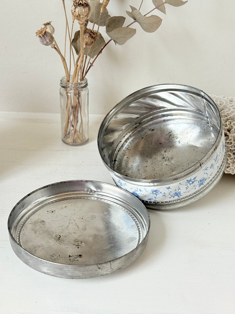 Vintage round shortbread tin with blue floral pattern on silver background, photographed on a white surface.