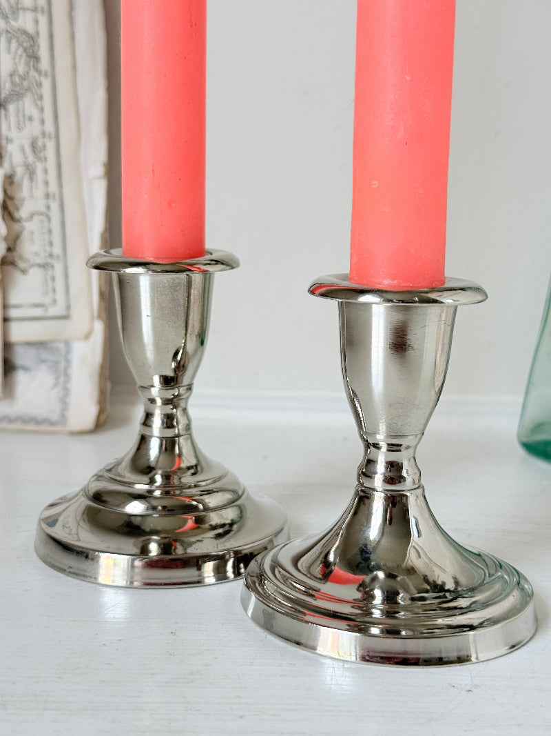 Pair of silver tone candlesticks with urn sconces and round stepped bases holding coral candles, photographed on a white surface