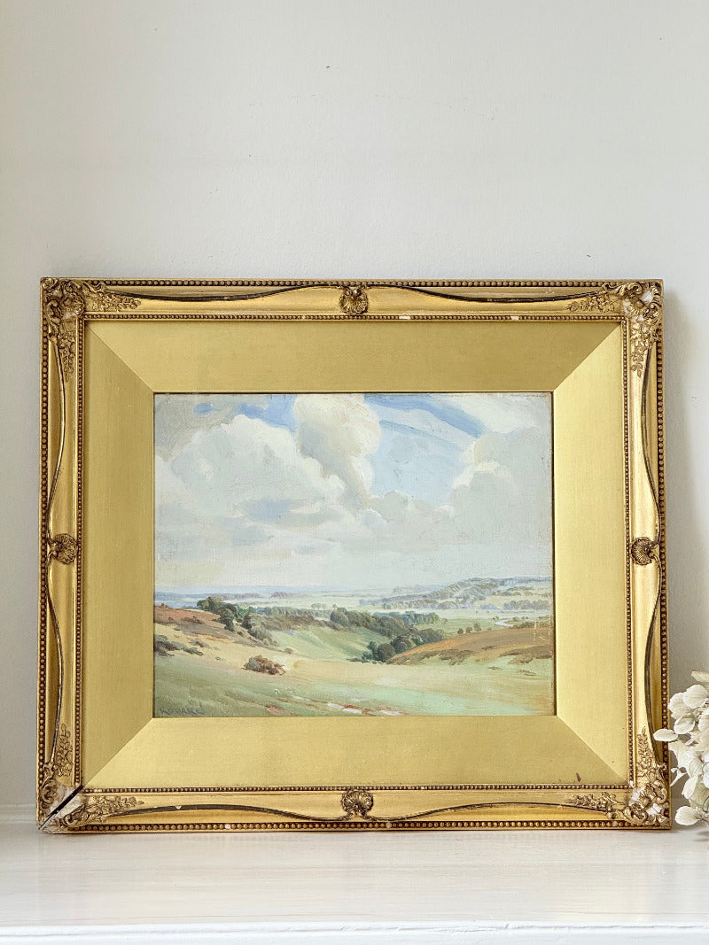 Framed oil painting of a rolling countryside landscape under a dramatic cloud-filled sky in an ornate gilt frame with wide mount.