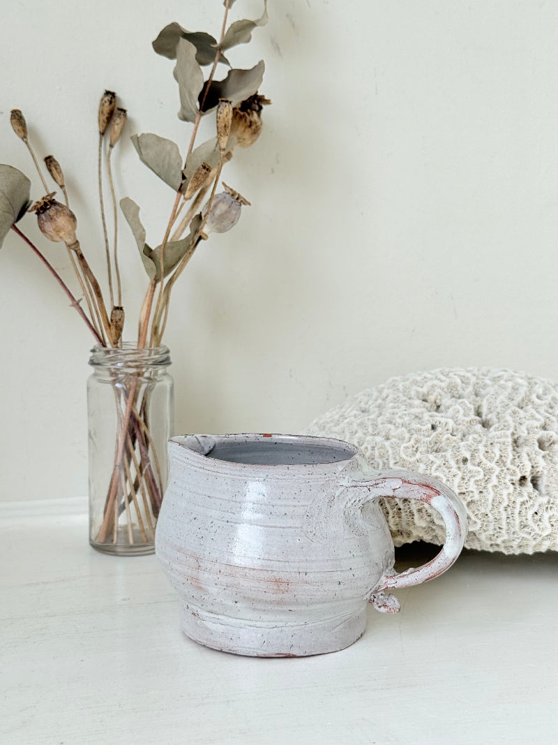 Studio pottery milk jug with soft pink and grey speckled glaze, photographed on a white surface