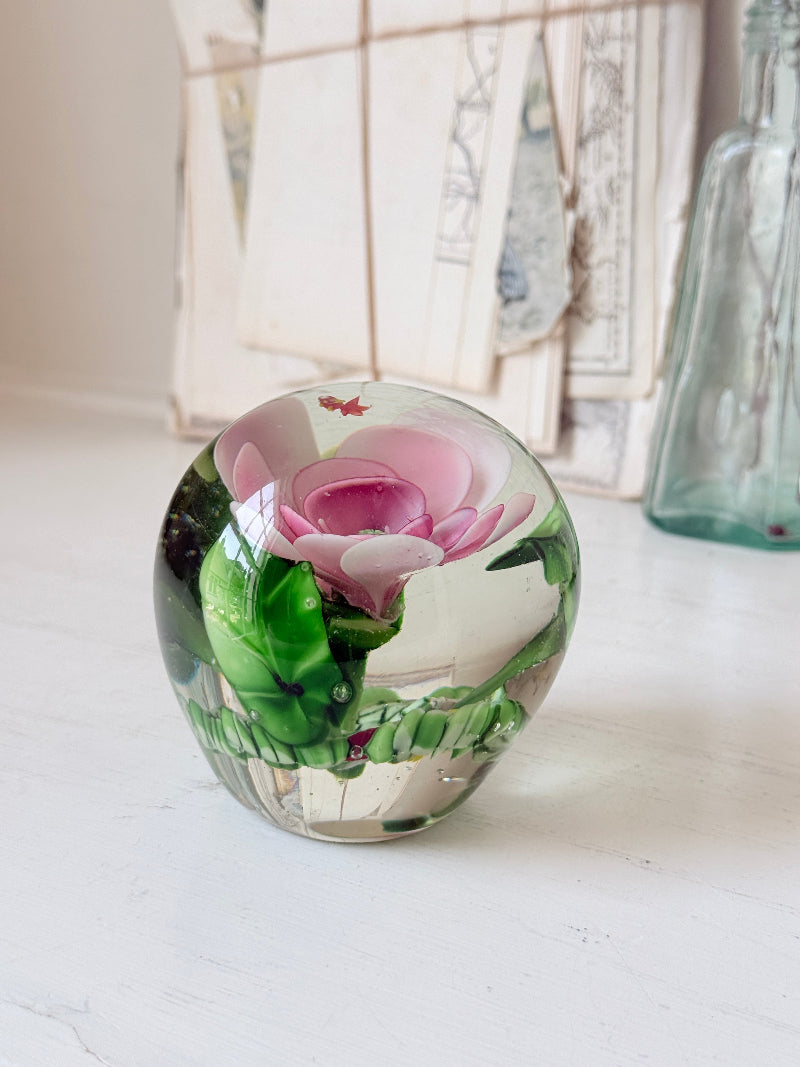 Large clear glass paperweight with a pink flower and green leaves inside, photographed on a white surface