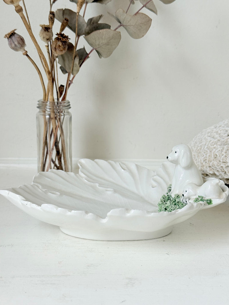 Italian ceramic dish featuring two white dogs on a sculpted leaf base, photographed on a white surface.