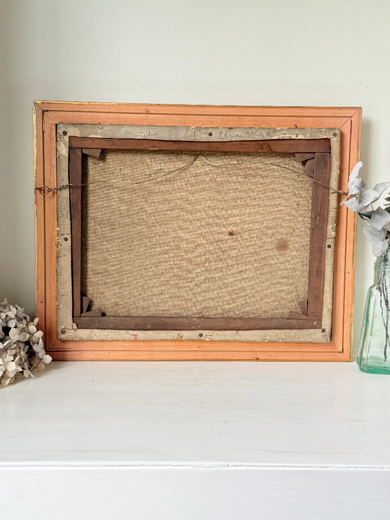An antique wooden picture frame, shown from the back, resting on a white surface against a light beige wall. The frame has a light orange-painted outer border, a darker brown inner frame, and a textured beige canvas backing.