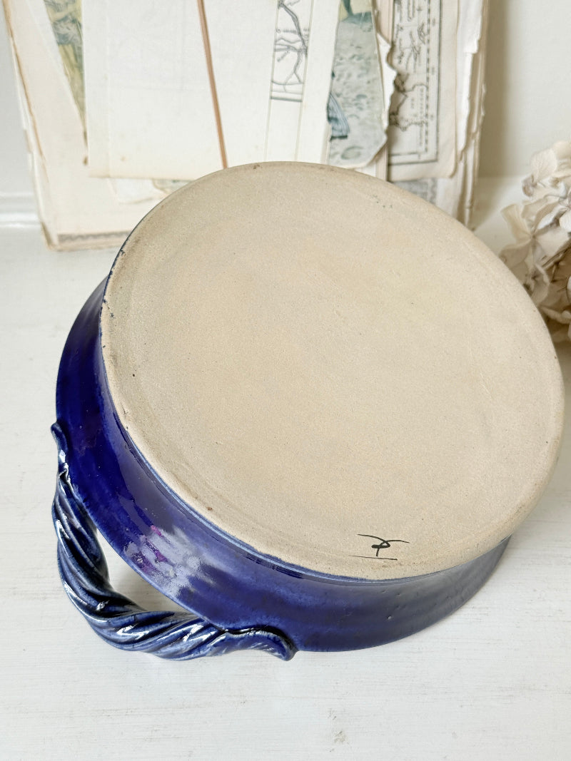 A handmade ceramic pot with a wide, unglazed beige clay top and a glossy cobalt blue glazed base and handle, resting on a white wooden surface.